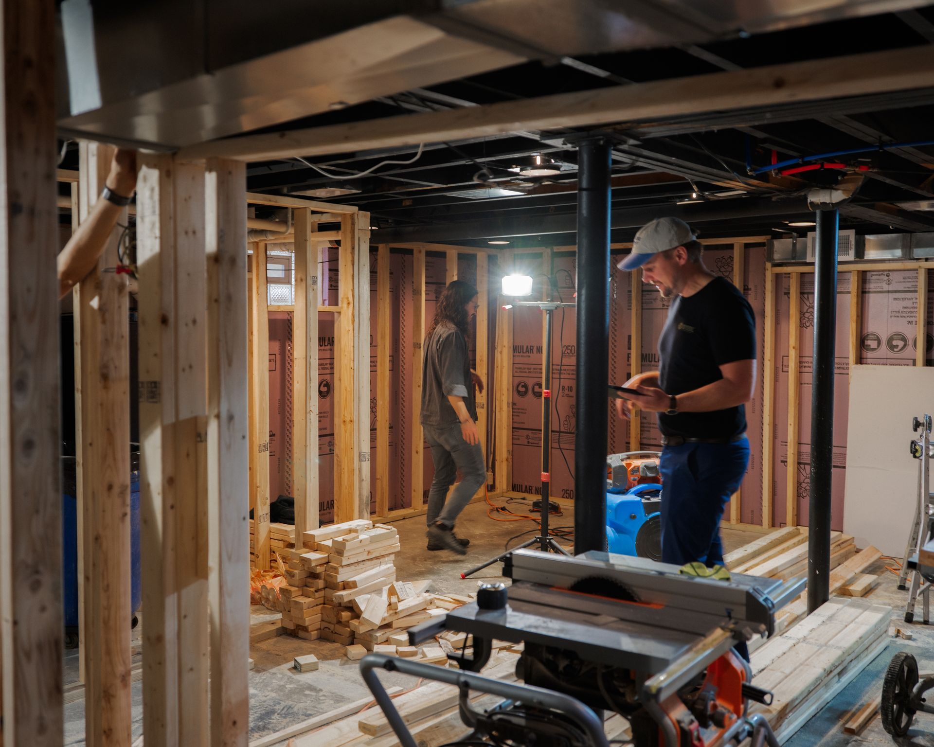 Two workers framing a basement interior during home renovation, with exposed studs and lumber stacks.