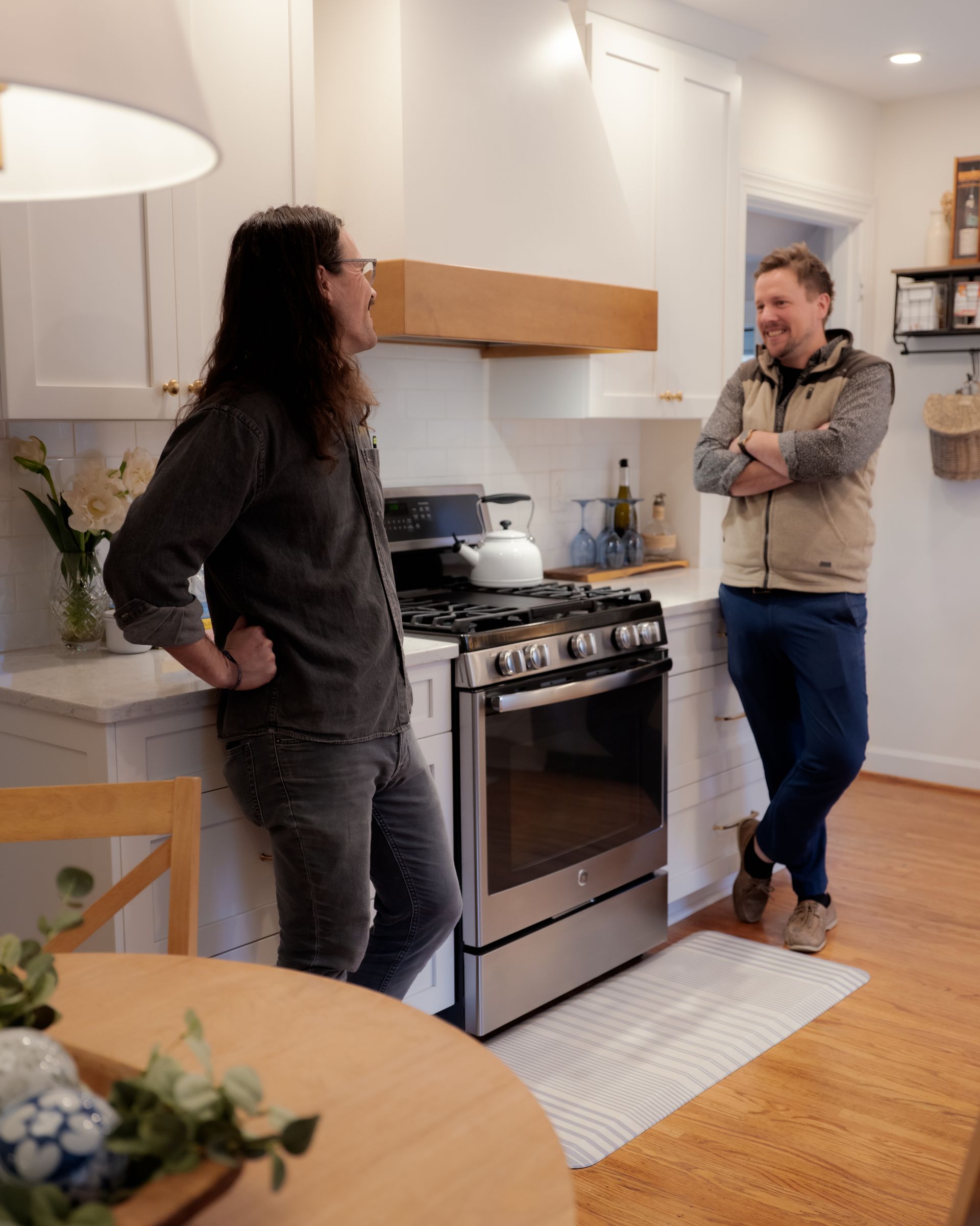 Two people talking in a bright kitchen, one leaning on a stove and the other standing nearby.