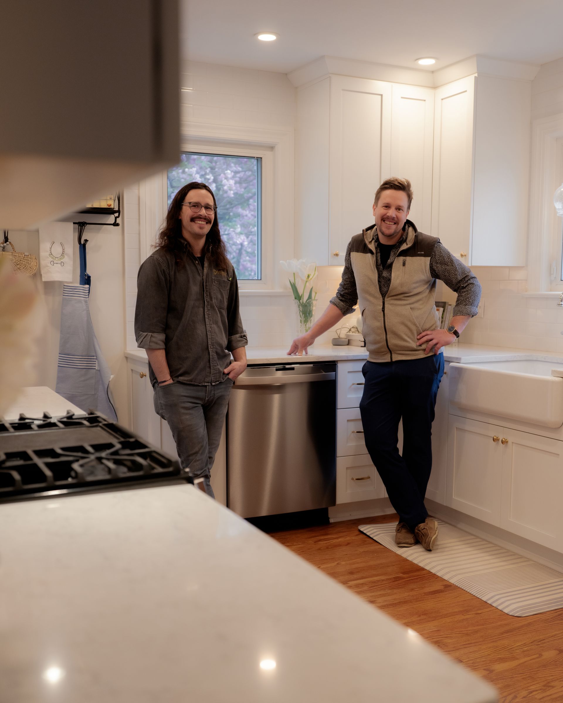 Two people standing in a bright white kitchen beside a stainless steel dishwasher, smiling