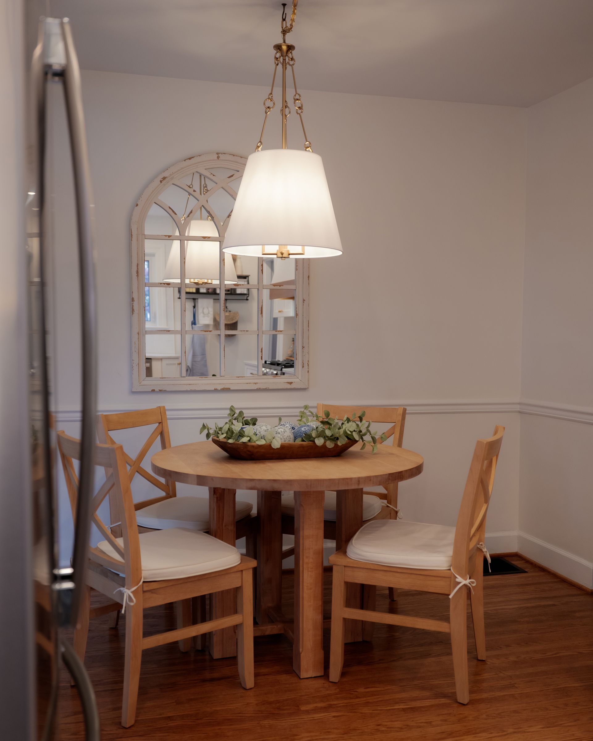 Dining nook with round wooden table, three chairs, hanging lamp, and wall mirror in a bright room
