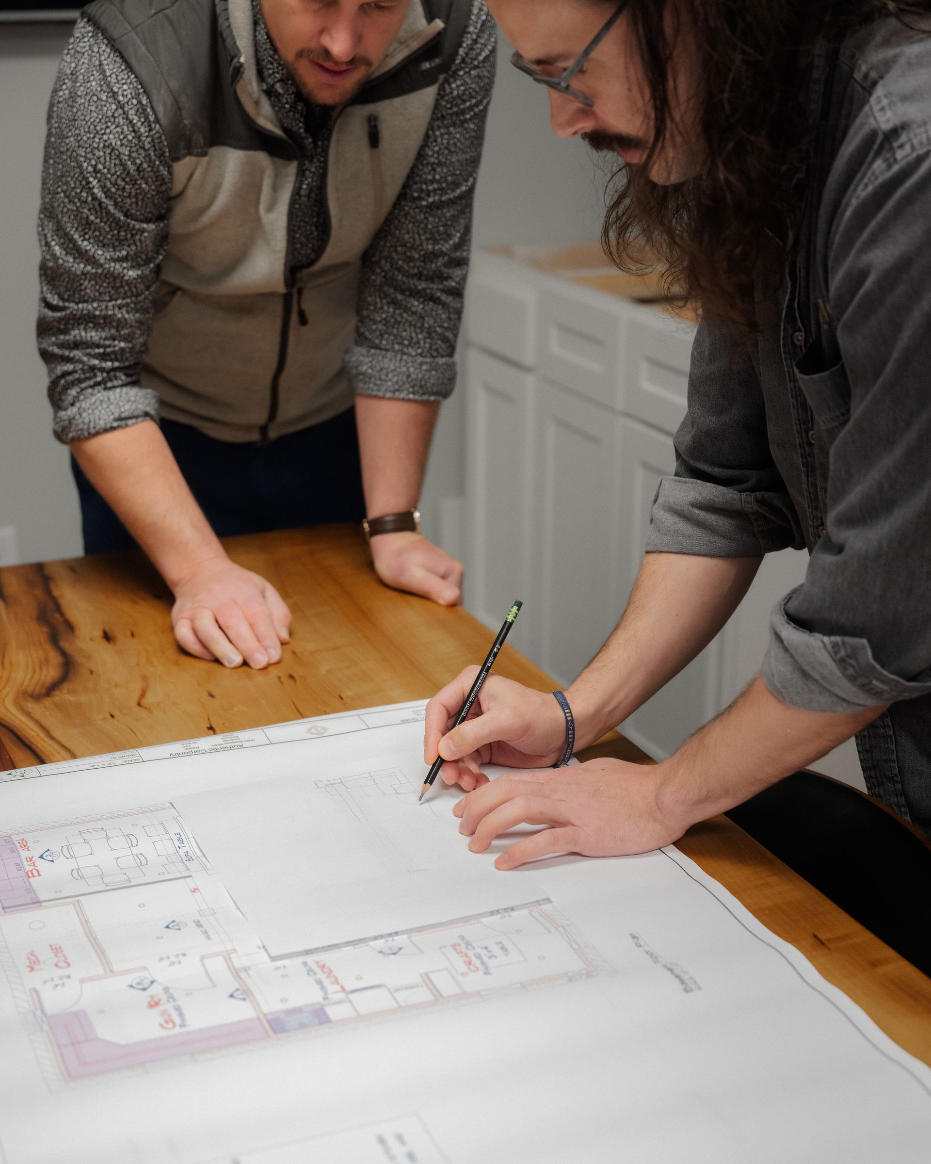 Two people reviewing a floor plan on a wooden table, one pointing with a pencil.