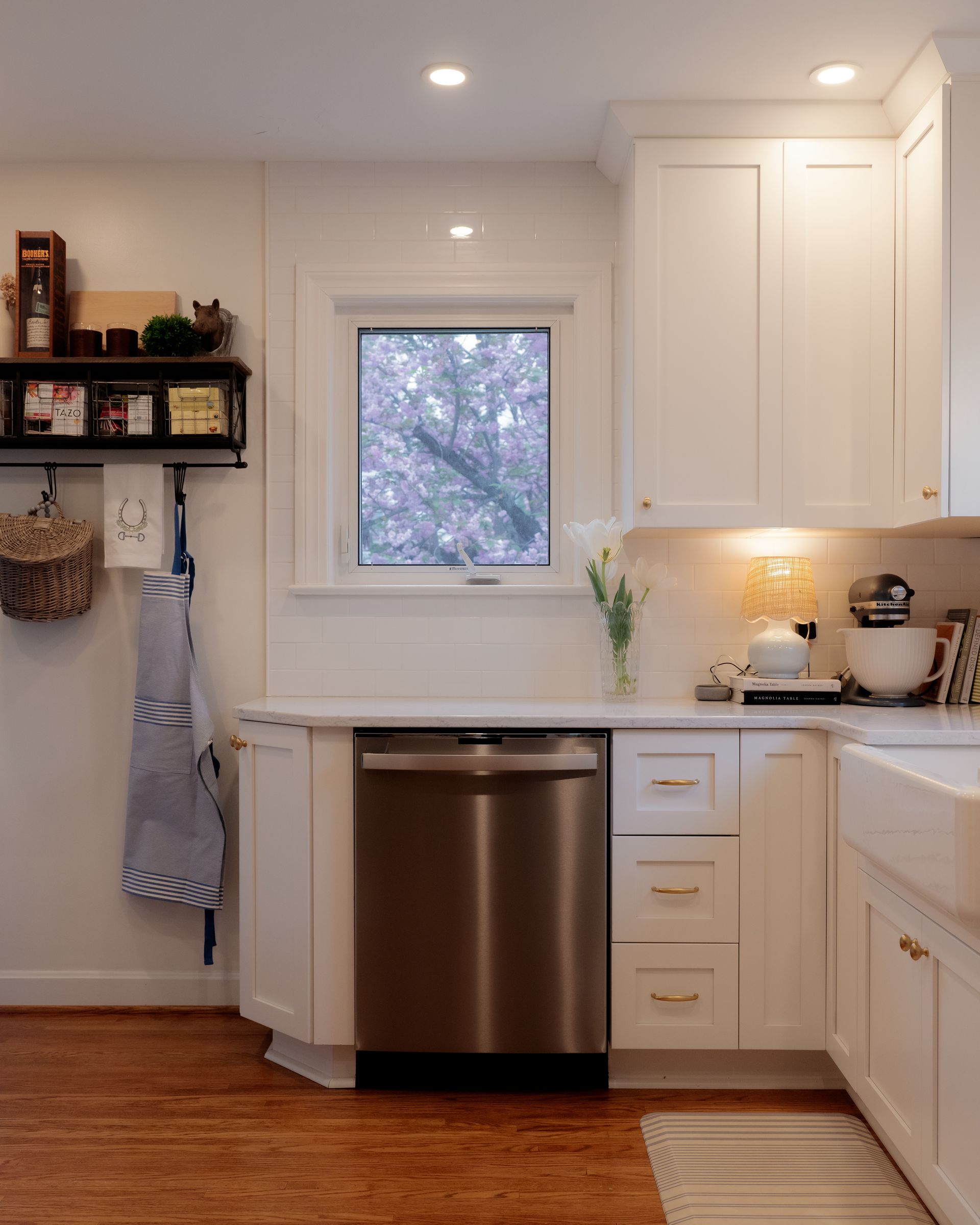 Bright white kitchen with stainless dishwasher, window over sink, and wood floor; shelves and cabinets line the walls.