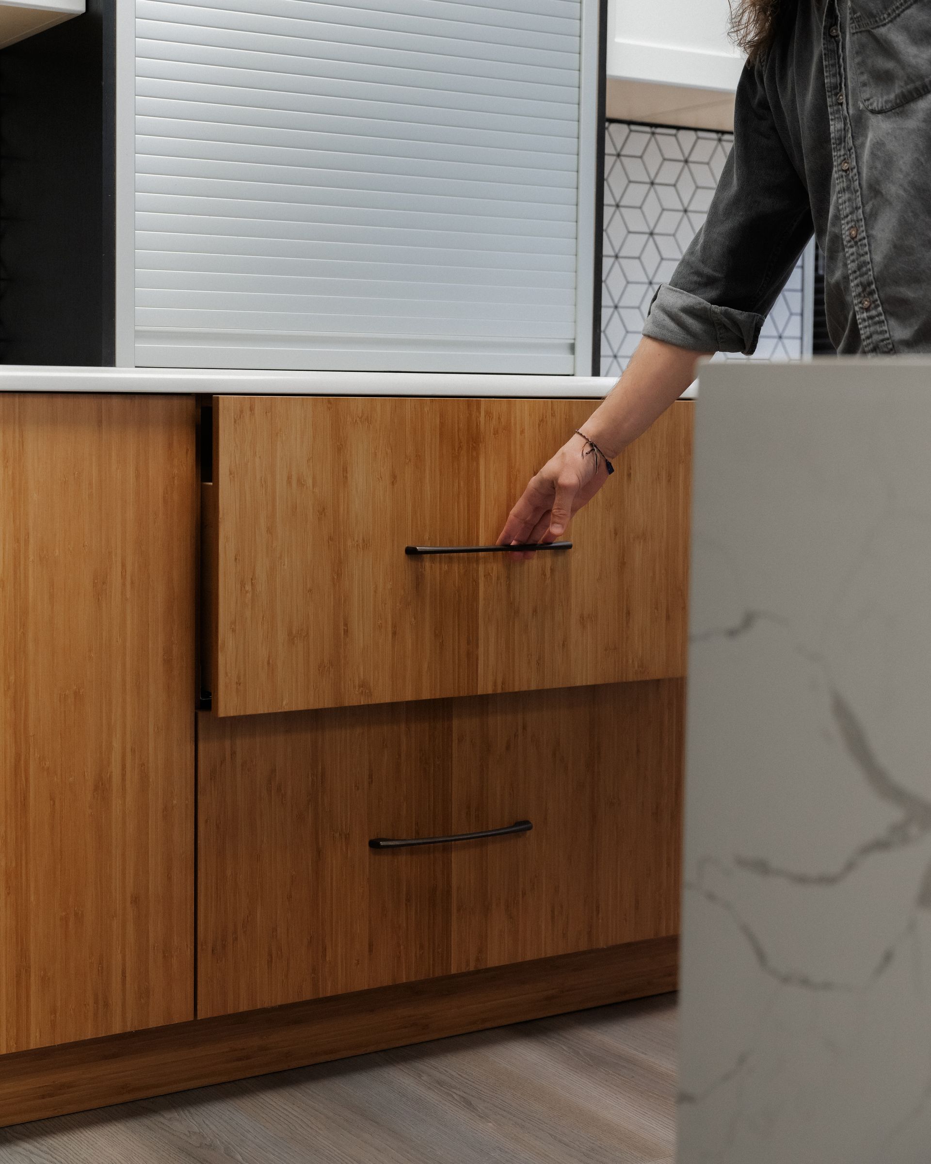 Hand reaching for a handle on a light wood kitchen cabinet beside a white countertop.