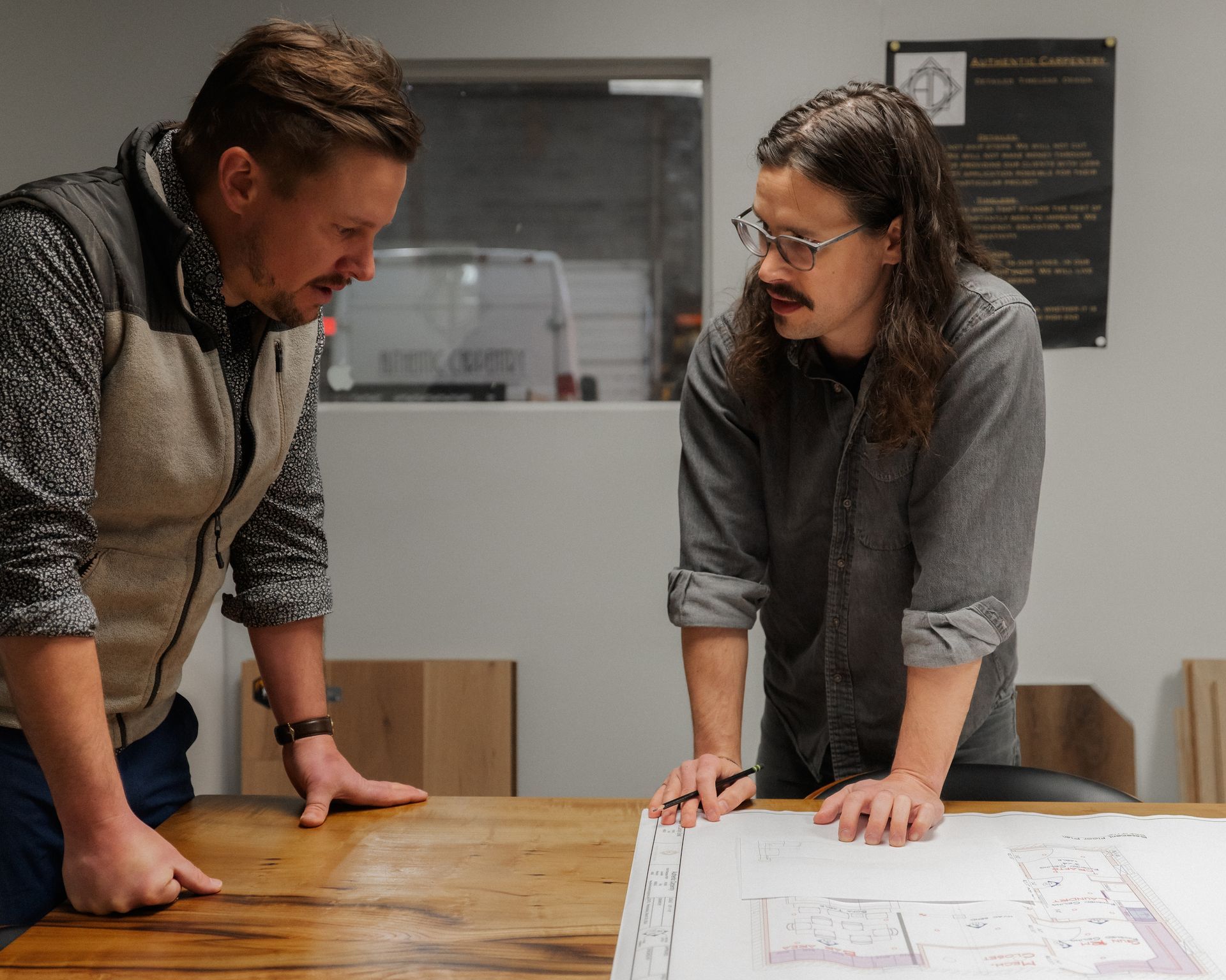 Two people leaning over a wooden table, reviewing plans in a bright office.