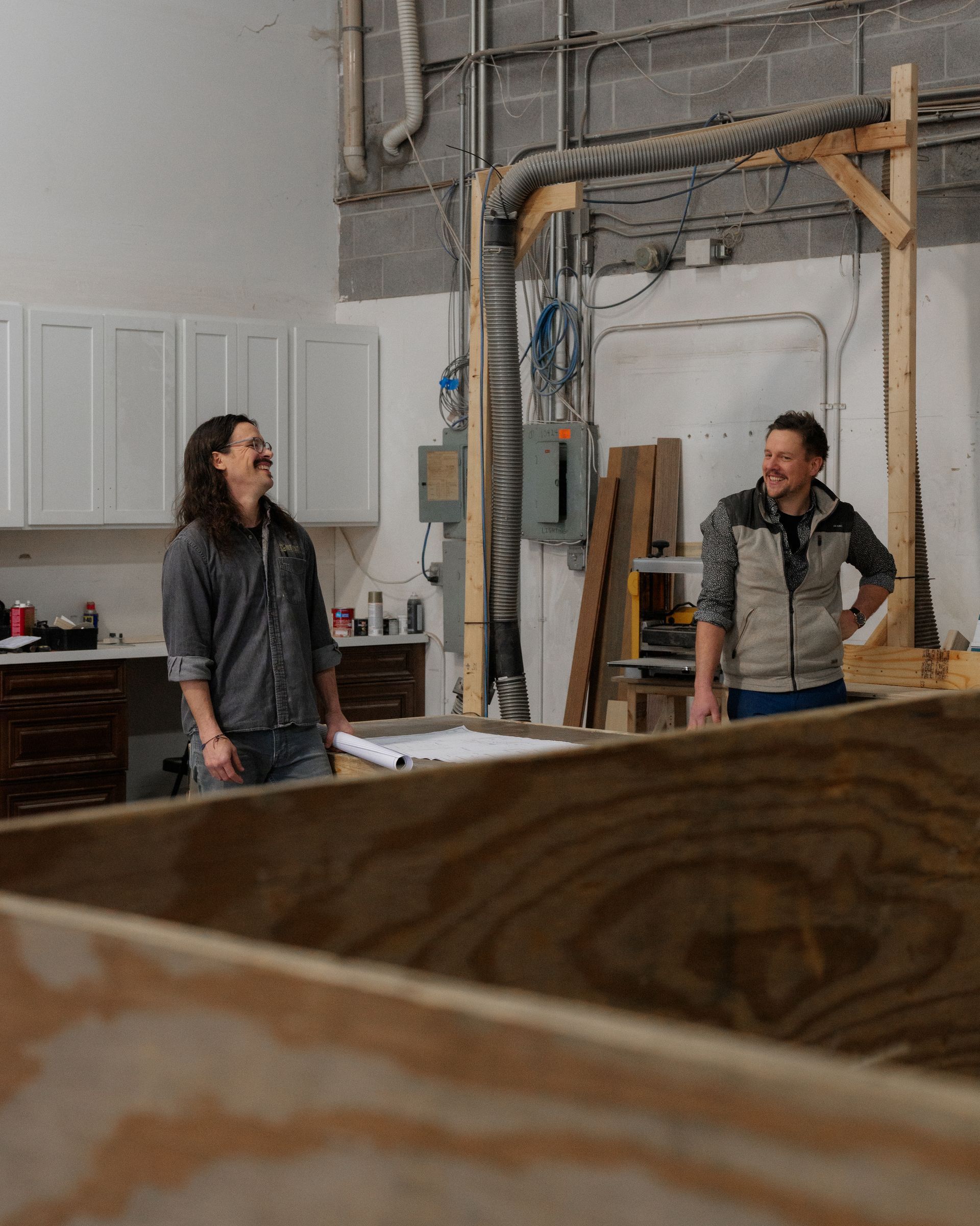 Two people stand in a workshop beside tools, cabinets, and stacked wood.