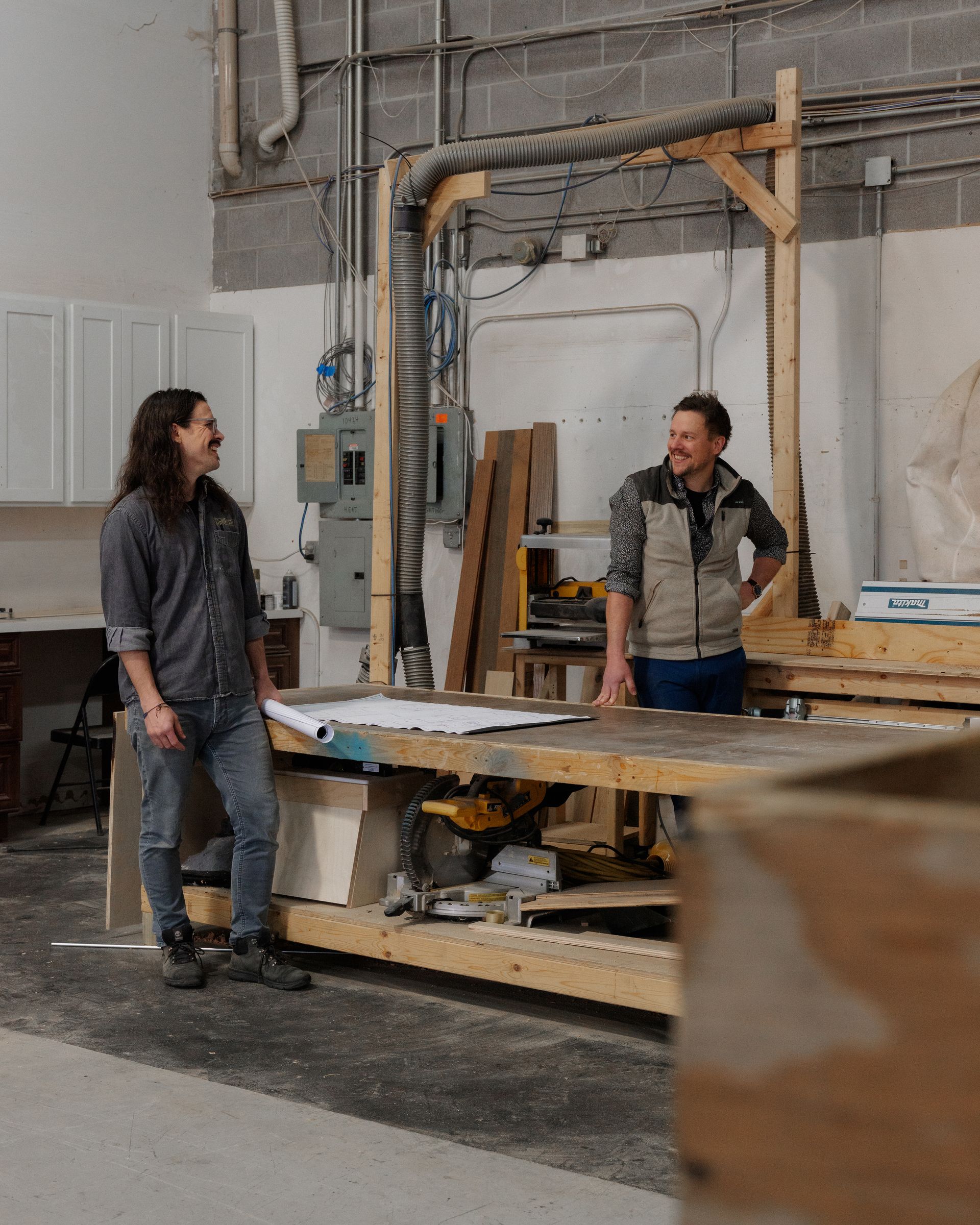 Two people standing and talking in a workshop beside a large wooden workbench and unfinished wall.