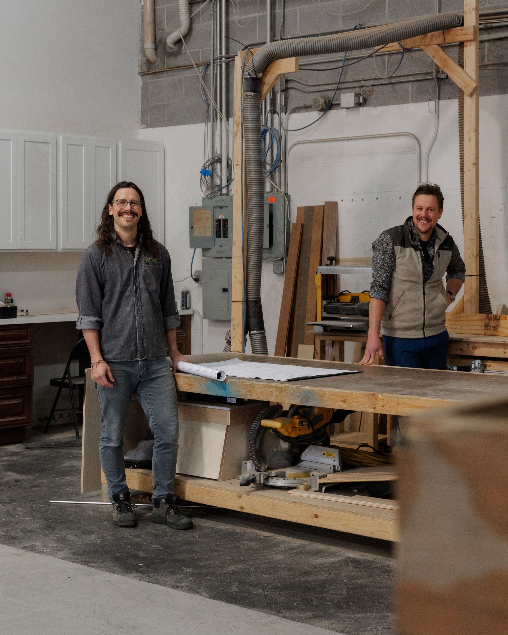 Two people smiling in a workshop beside a large workbench and tools
