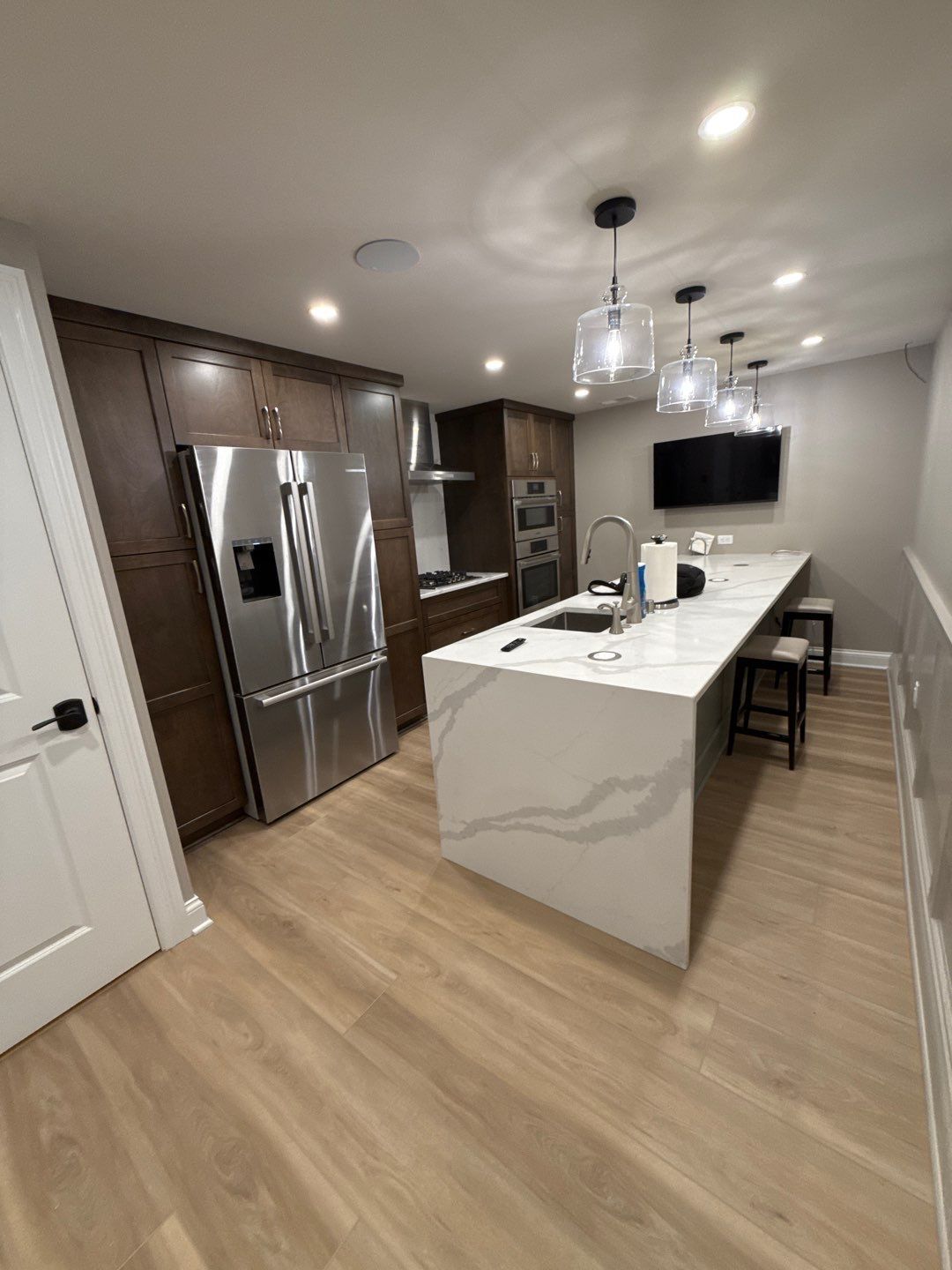 Modern kitchen with white island, stainless steel fridge, pendant lights, and wall-mounted TV
