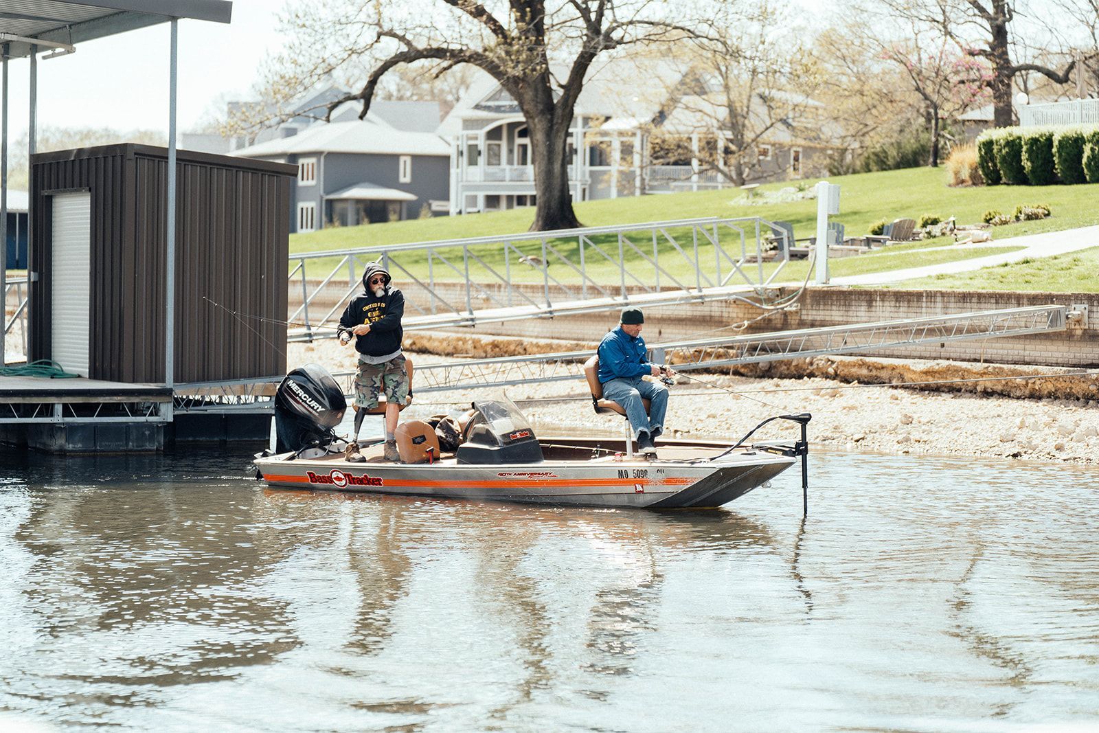 Two men fishing from a boat near a dock; houses and trees in the background.