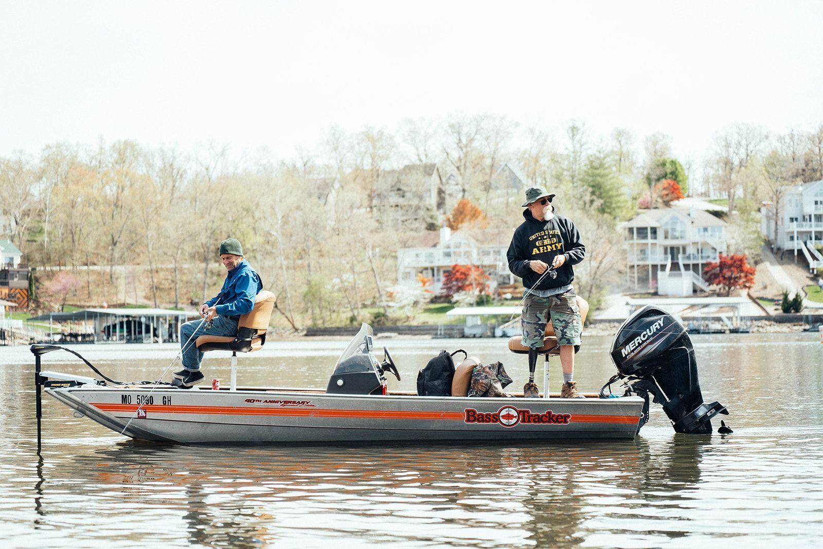Two men fishing from a Tracker boat on a lake, houses in the background.