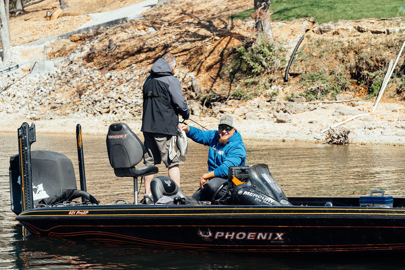 Two people on a Phoenix boat, one holding a fish, near a rocky shore on a sunny day.