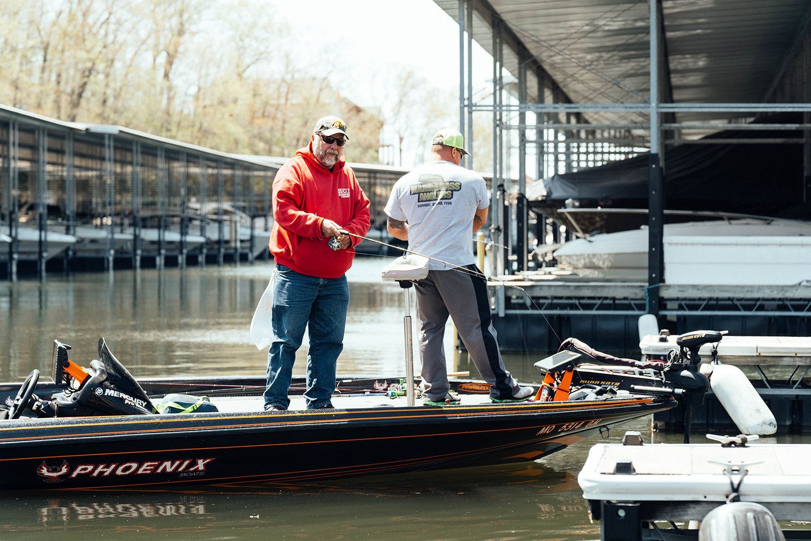 Two men fishing from a boat near a dock; one in red hoodie, the other in a white tee.