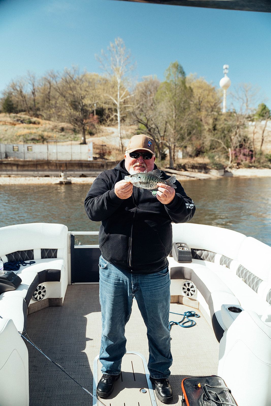 Man on boat holding up a fish. He wears a cap, jacket and jeans, with a calm lake and trees in the background.