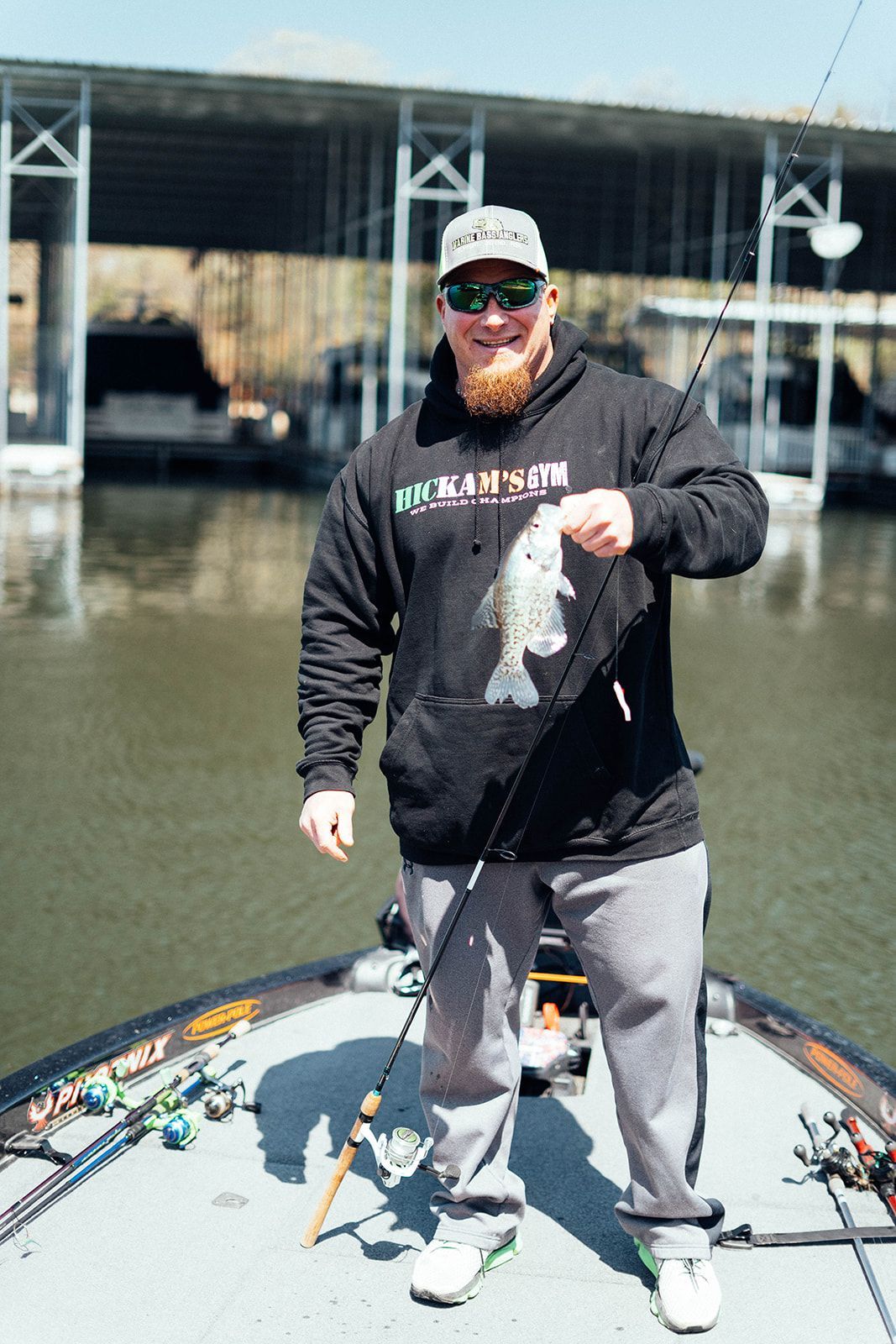 Man on boat holds a fish; docks and lake in the background. He wears a hoodie, hat and smiles.