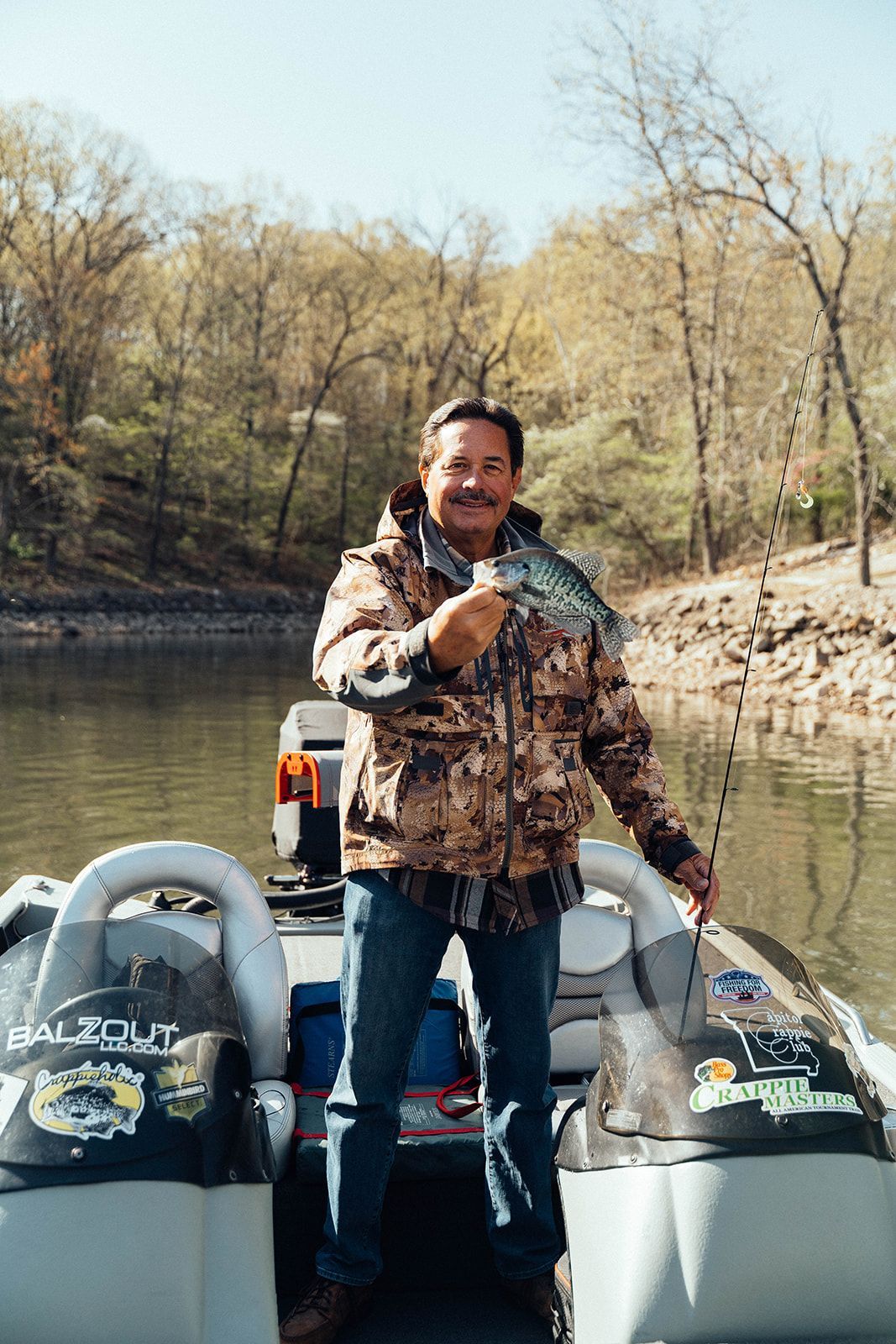 Man on a boat holding a fish, smiling. He wears a camo jacket and jeans. Lakeside background.