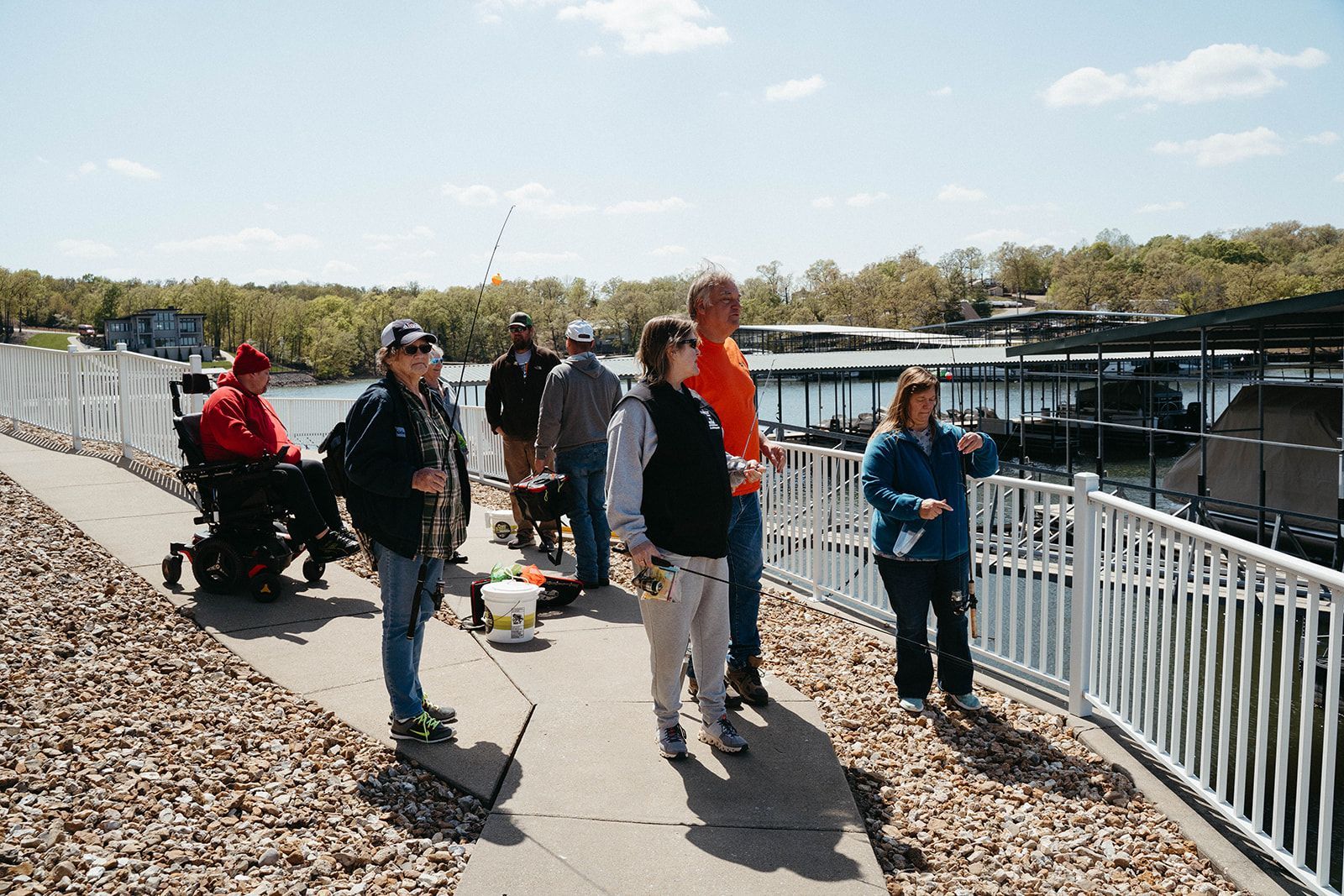 People fishing from a pier, one in a wheelchair. Sunny day, boats docked in background.