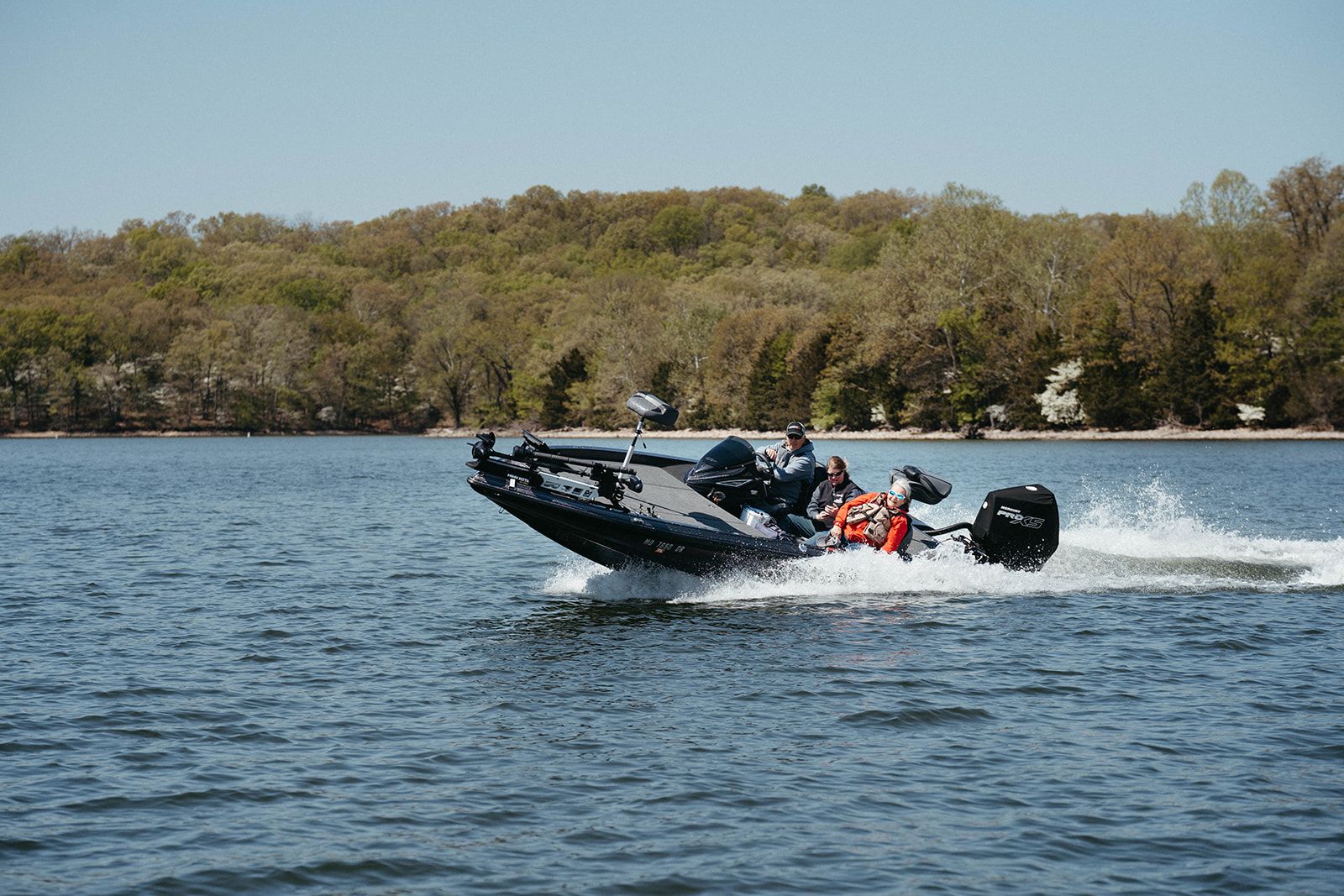 A dark fishing boat speeding across a lake with two people in it, trees in background.