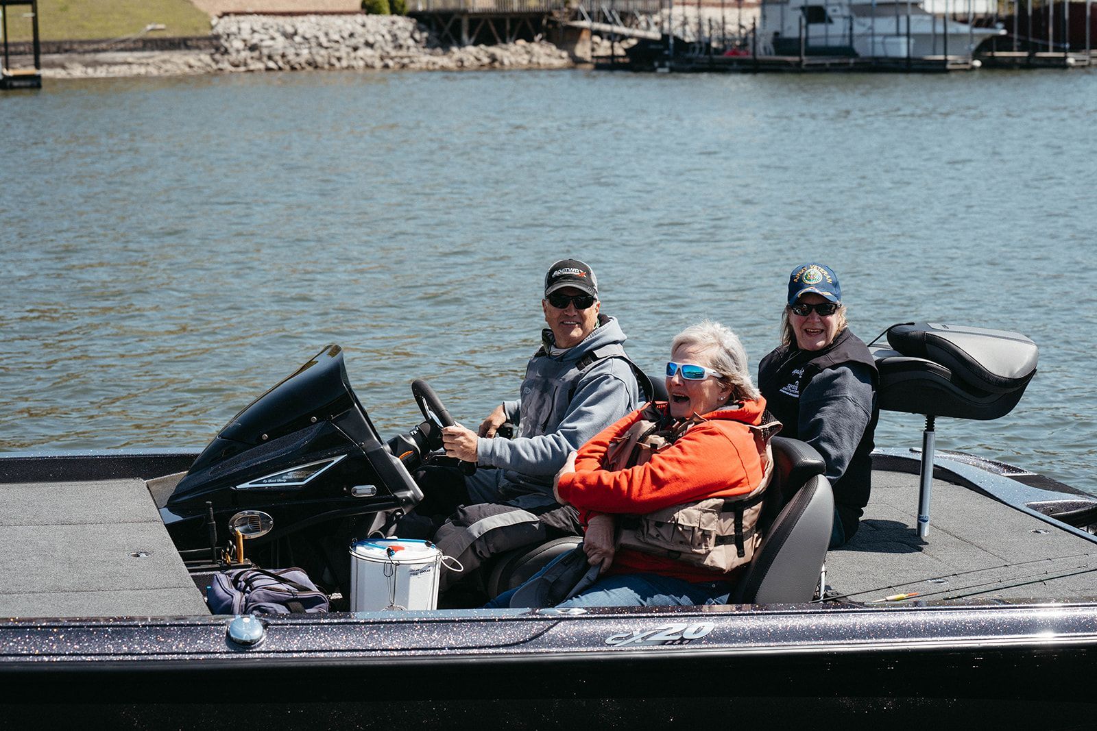 Three people smile aboard a boat on a lake; one drives, one wears an orange jacket.
