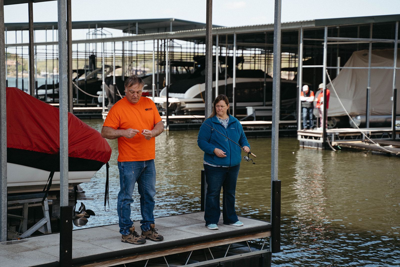 Man and woman fishing off a dock with boats docked in the background; the man wears orange, the woman wears blue.