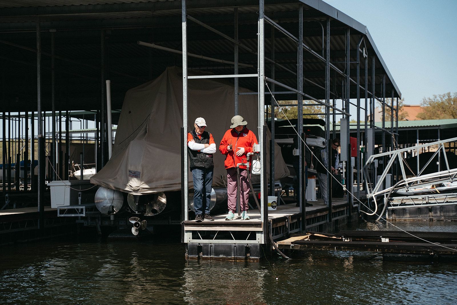 Two people standing on a dock near a boat under a covered structure at a marina.