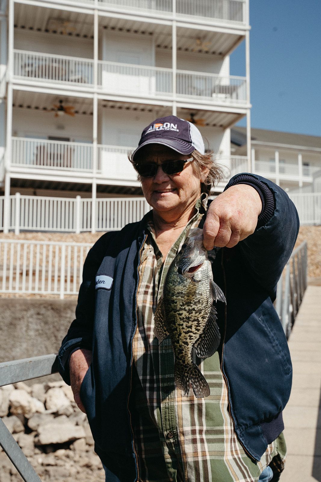 Woman in cap holding up a crappie fish on a dock in front of a white building.