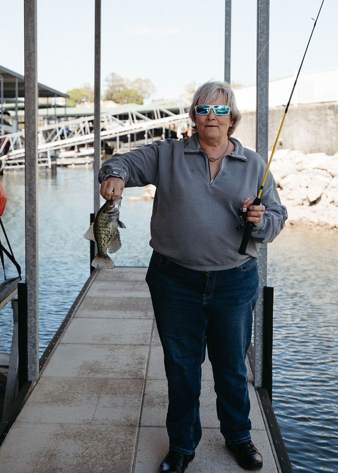 Woman on dock holds up a spotted fish; fishing rod in hand. Waterfront setting, blue water.
