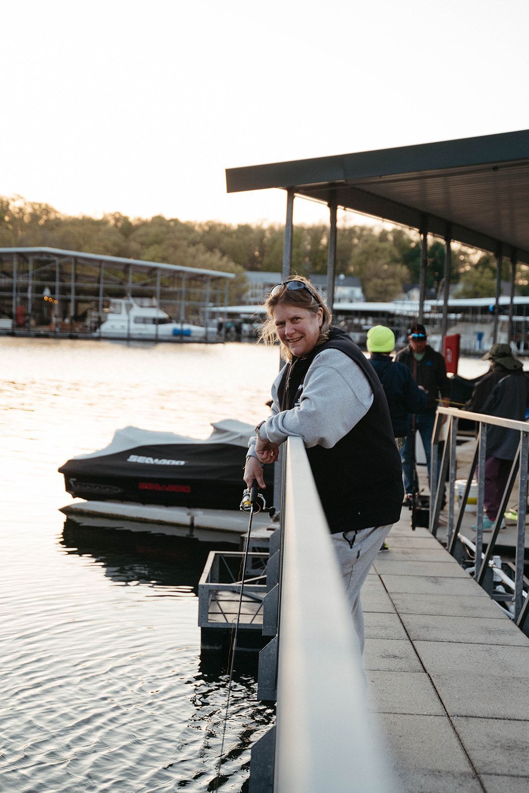 Woman smiling on a dock, leaning on railing near a jet ski and water. Sunset at a marina.