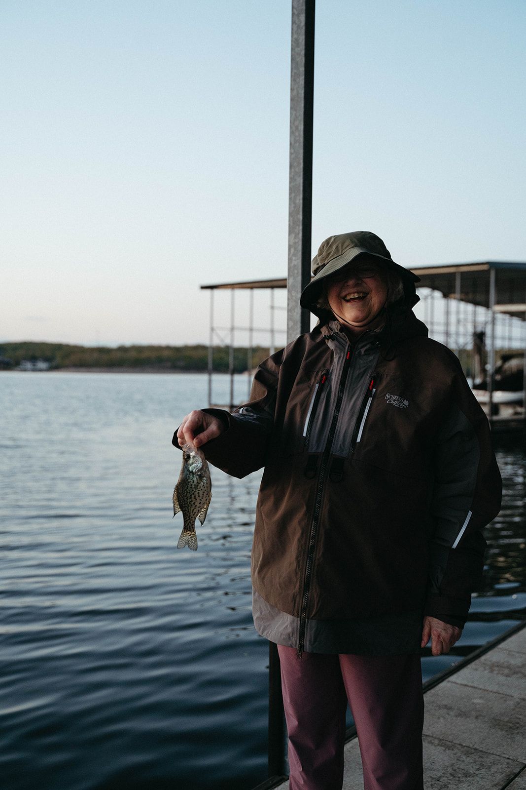Person on dock holds up a small fish, smiling. Wearing a hat and jacket; lake and sky in the background.
