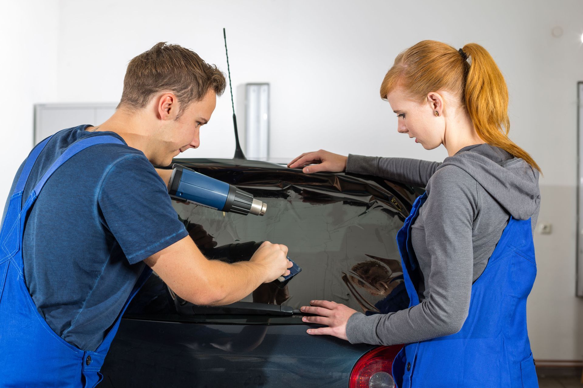 Two people applying tinted film to a car's rear window. One uses a heat gun, the other smooths the film.