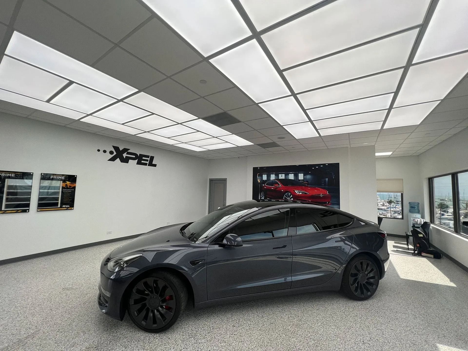Dark gray Tesla Model 3 in a car detailing shop with XPEL branding on the wall.