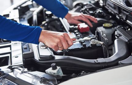 Person checking a car engine with hands inside the open hood, using a tool in a garage.