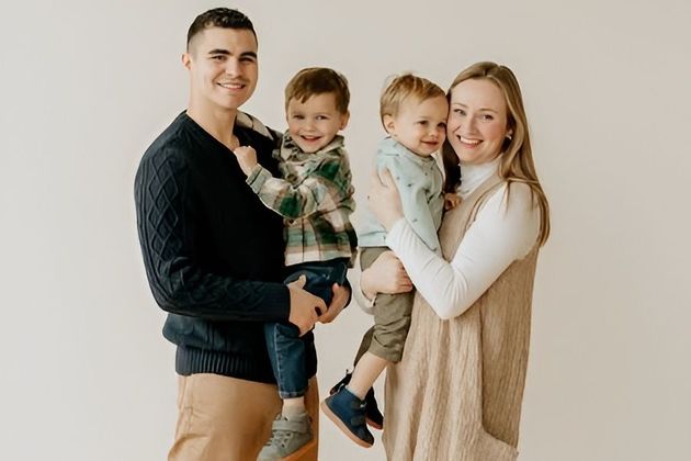 A smiling family of four posing against a plain background, with two parents each holding a small child.