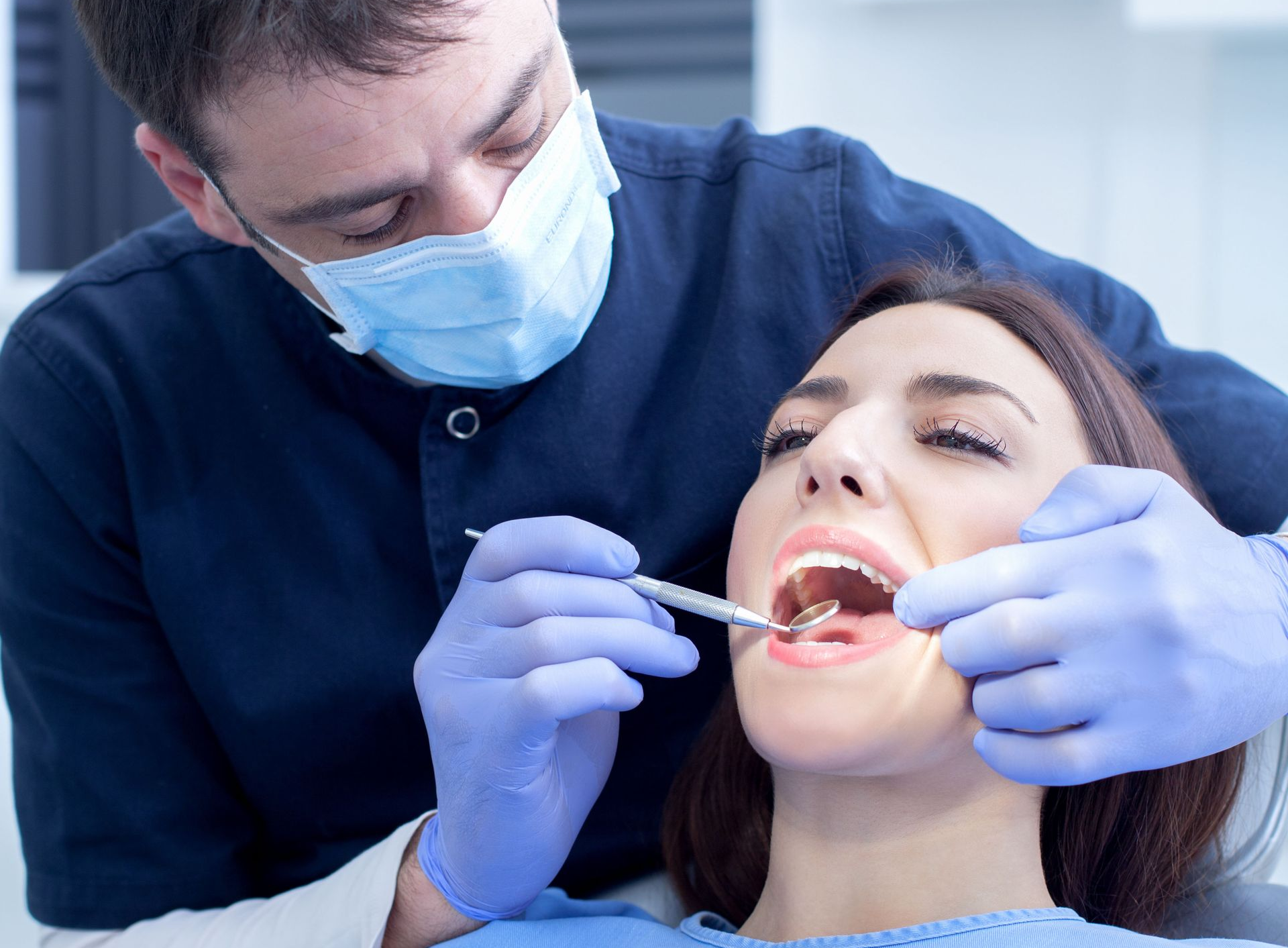 Dentist examining a patient's mouth with tools in a dental office. The patient is sitting, mouth open.