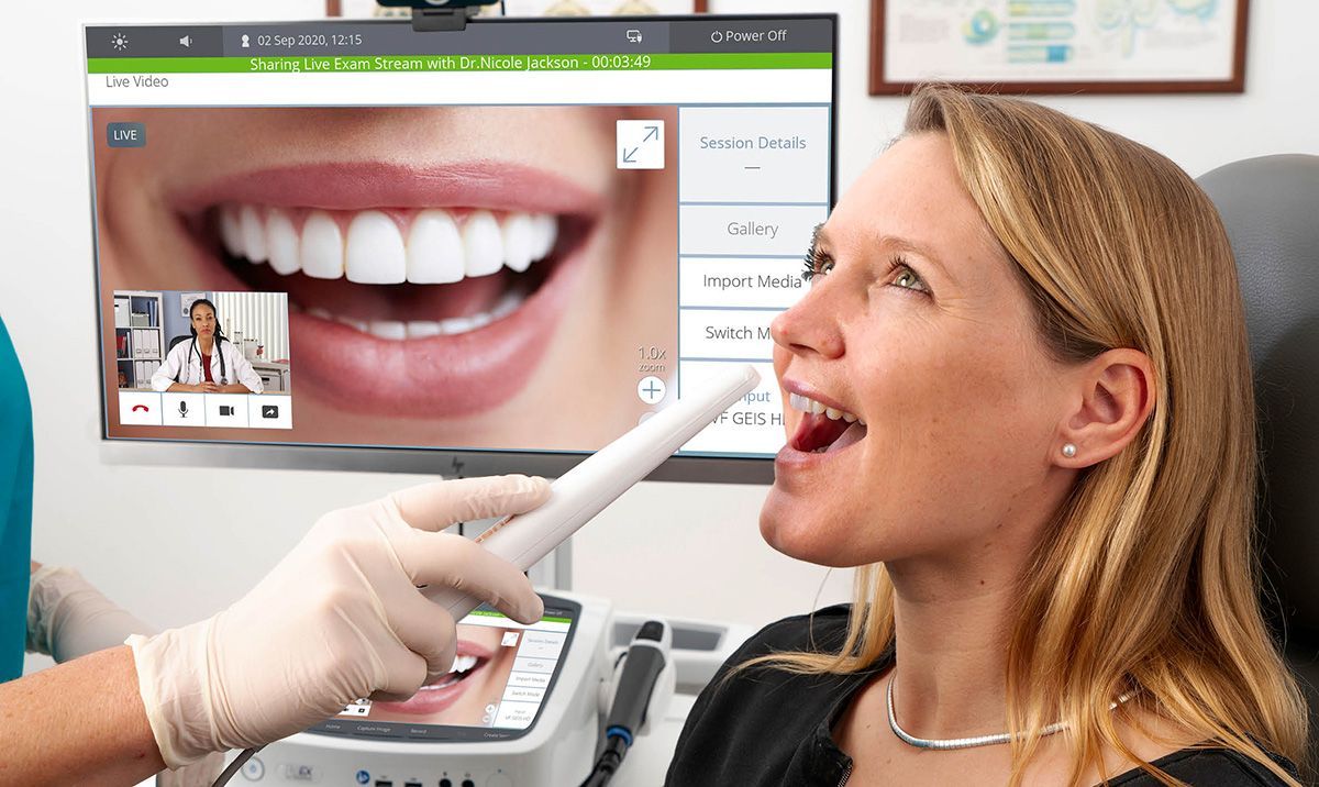 A dentist uses an intraoral camera on a patient, with the dental imagery displayed on a screen in a clinical setting.