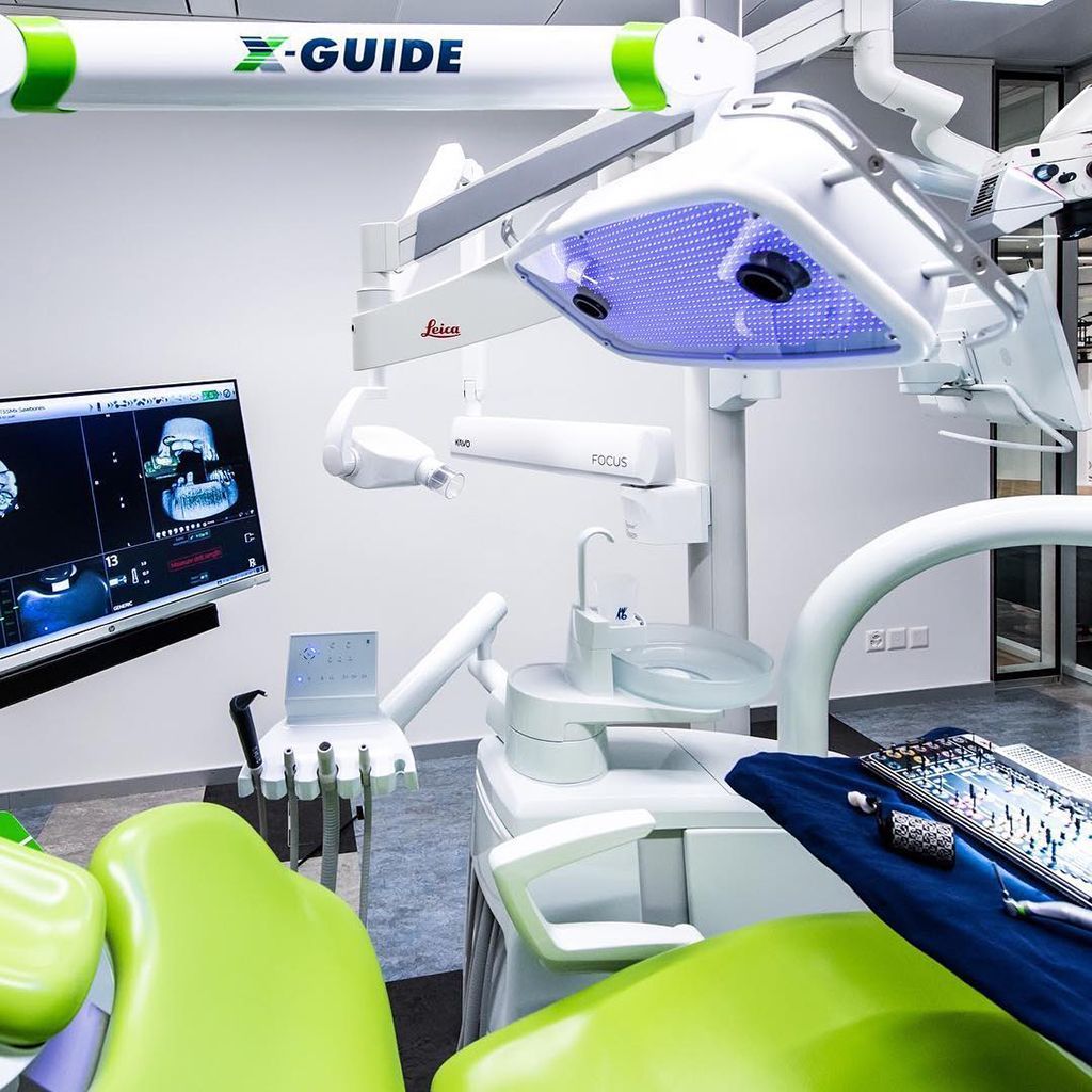 A modern dental office featuring a green patient chair, X-Guide surgical equipment, and a monitor displaying dental scans.