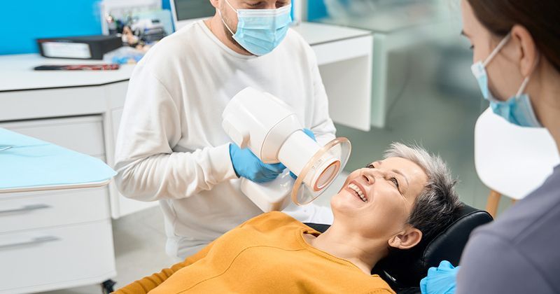 A dental professional uses a handheld X-ray device on a patient sitting in a clinical office setting.