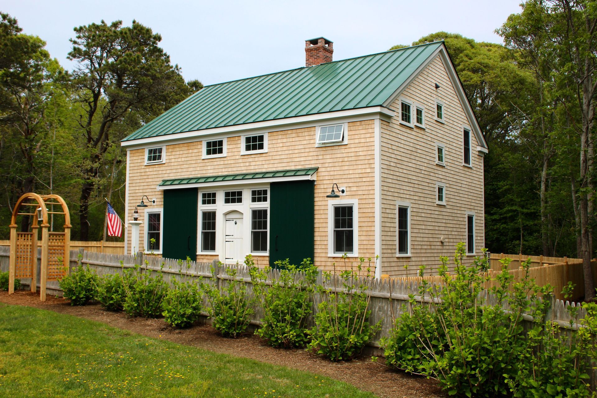 Two-story beige house with green roof and shutters, surrounded by a wooden fence and greenery