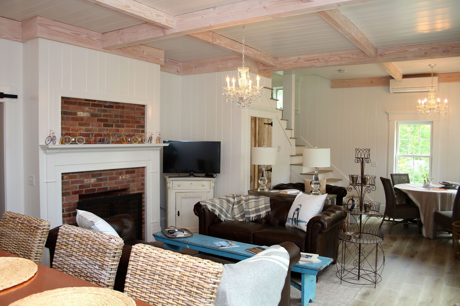 Living room with fireplace, leather couch, chandelier, and dining area; white walls, wood beams.