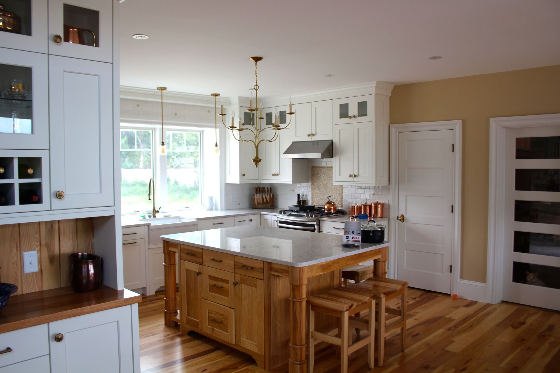 Spacious kitchen with white cabinets, light wood island and floors. Beige walls, natural light, and a gold chandelier