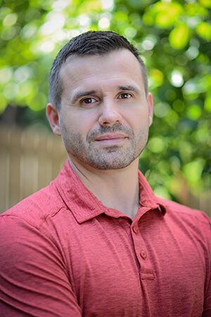 A man with a beard is wearing a red shirt and standing in front of a fence.