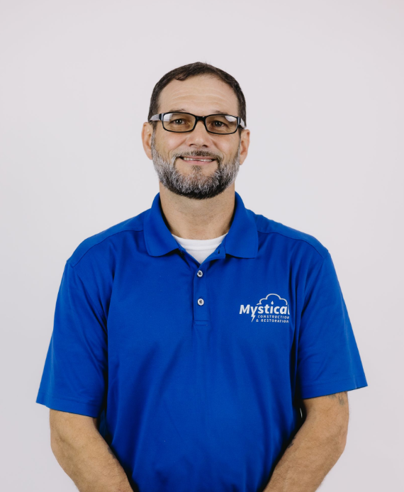 Man in blue polo shirt and glasses, standing against a plain light background, smiling gently