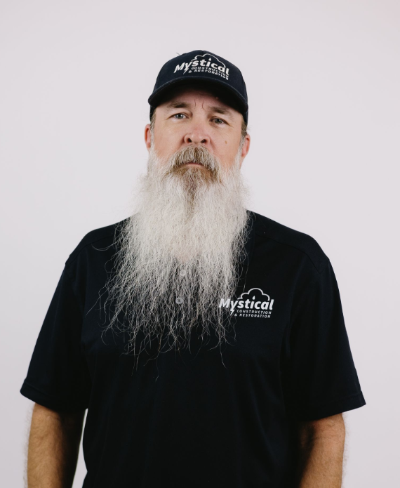 Man in black cap and black T-shirt with long gray beard against a light background