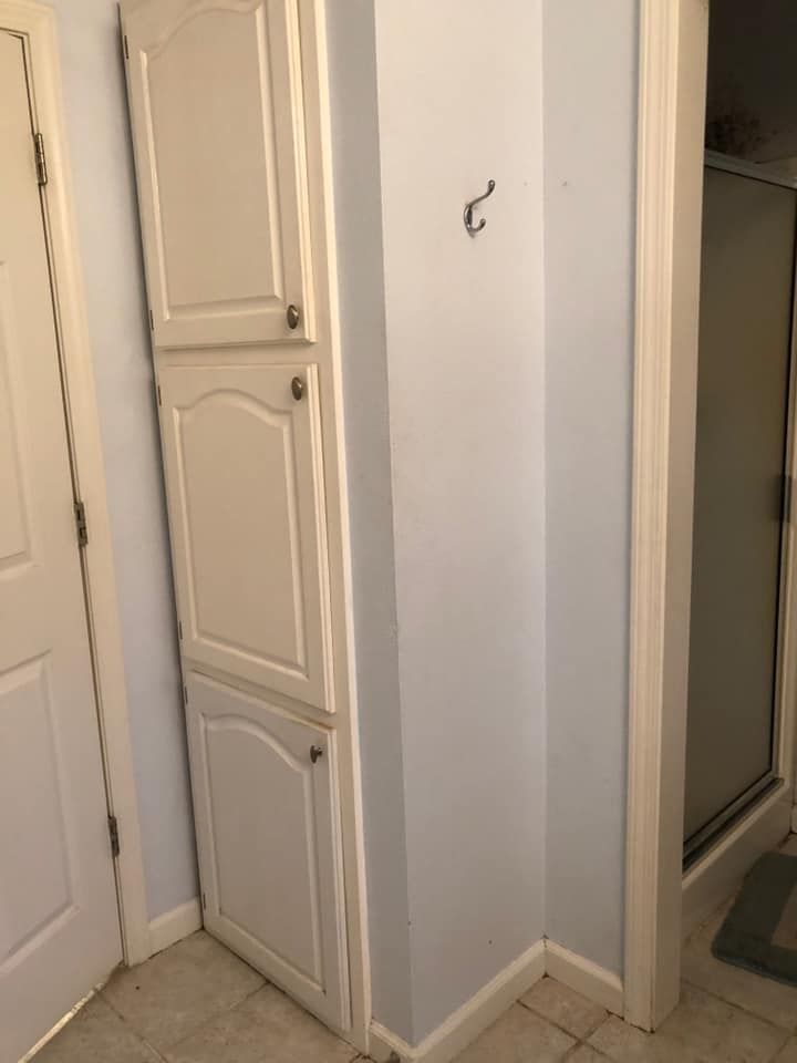 White storage cabinet in bathroom corner, next to light blue wall and shower.
