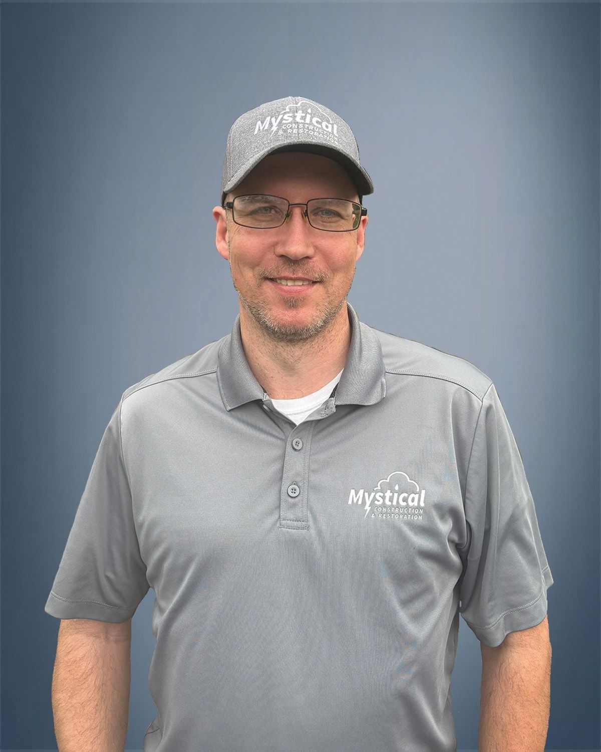 Man wearing glasses and a hat with "Mystical" logo on a grey shirt, against a blue background.