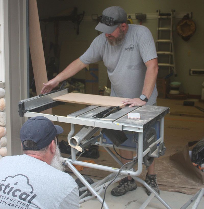 Two construction workers using a table saw to cut wood inside a garage.