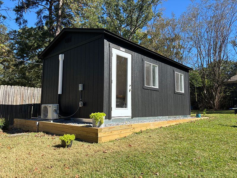 Black tiny house with white trim, door, and windows, set on a wooden base in a grassy yard.
