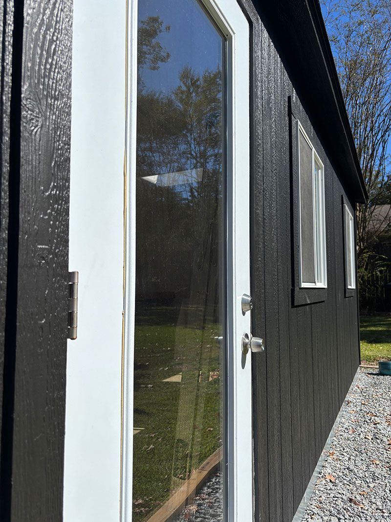 Black-sided building with white-framed glass door and windows. Gravel path next to the building.