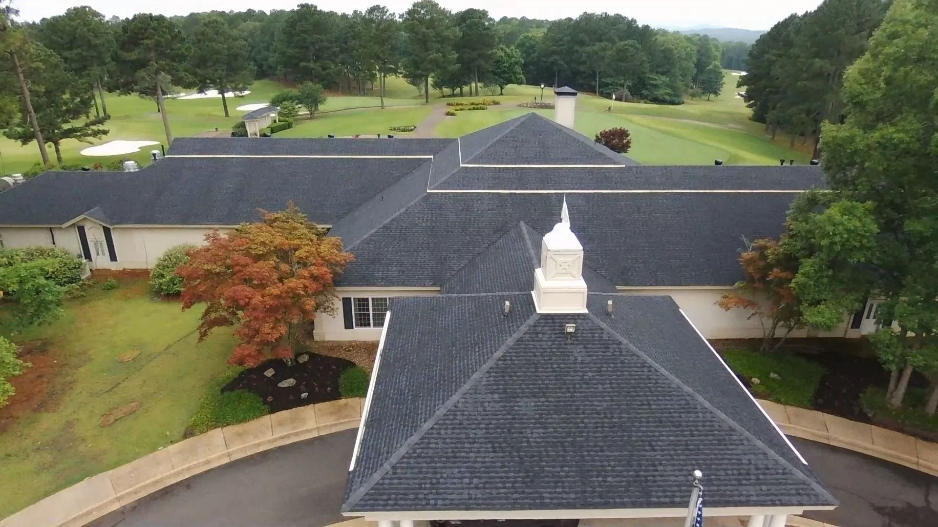 A building with a dark roof and white trim surrounded by green grass and trees, likely a golf course clubhouse.