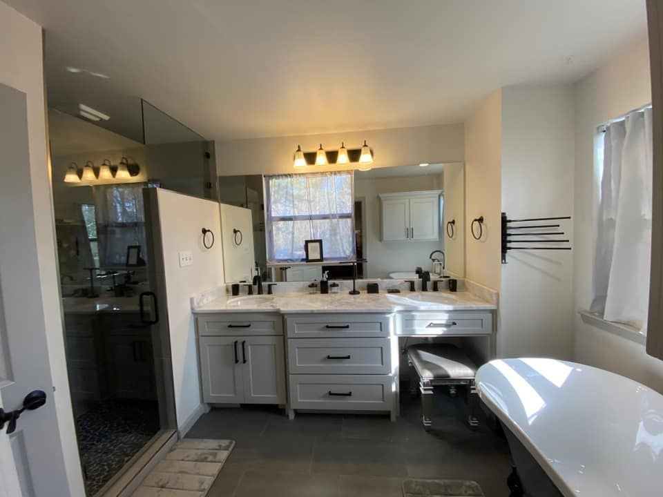 Bathroom with double vanity, shower, and freestanding tub. Gray cabinets, black fixtures, and a large mirror.