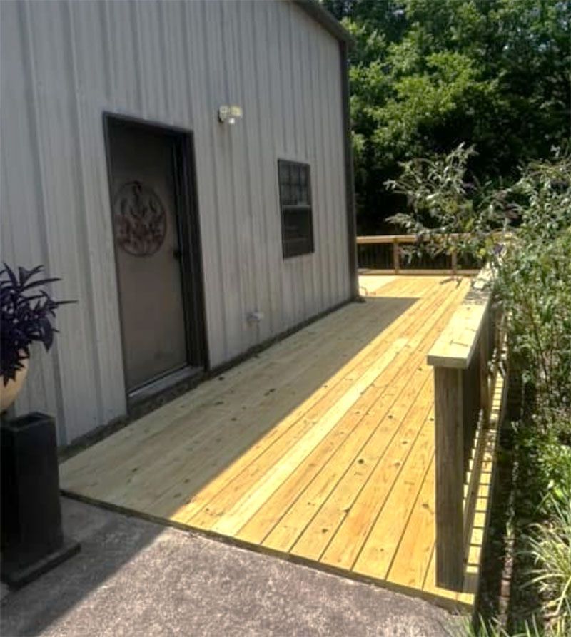 A wooden deck with a handrail extends from a building's door and window. Bright sunlight.